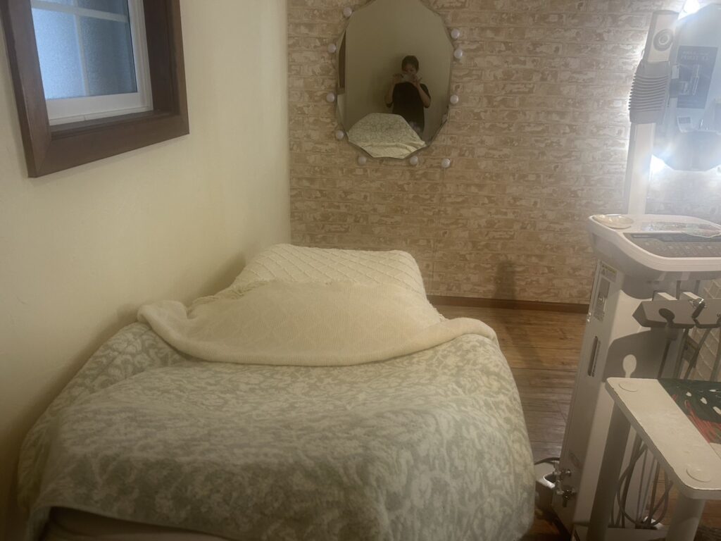 A cozy facial treatment room featuring a massage bed draped with a soft blanket, a mirror adorned with lights, and a facial treatment machine.